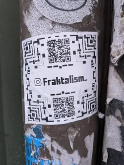 Street sticker A rectangular white sticker adhered to a dark, textured pole, featuring two prominent black and white QR codes—one at the top and one at the bottom. Centered between the QR codes is the Instagram logo followed by the text 'Fraktalism_'. The sticker shows signs of wear, with slight creases and dirt, indicating its outdoor placement. It is surrounded by remnants of other torn and faded papers.