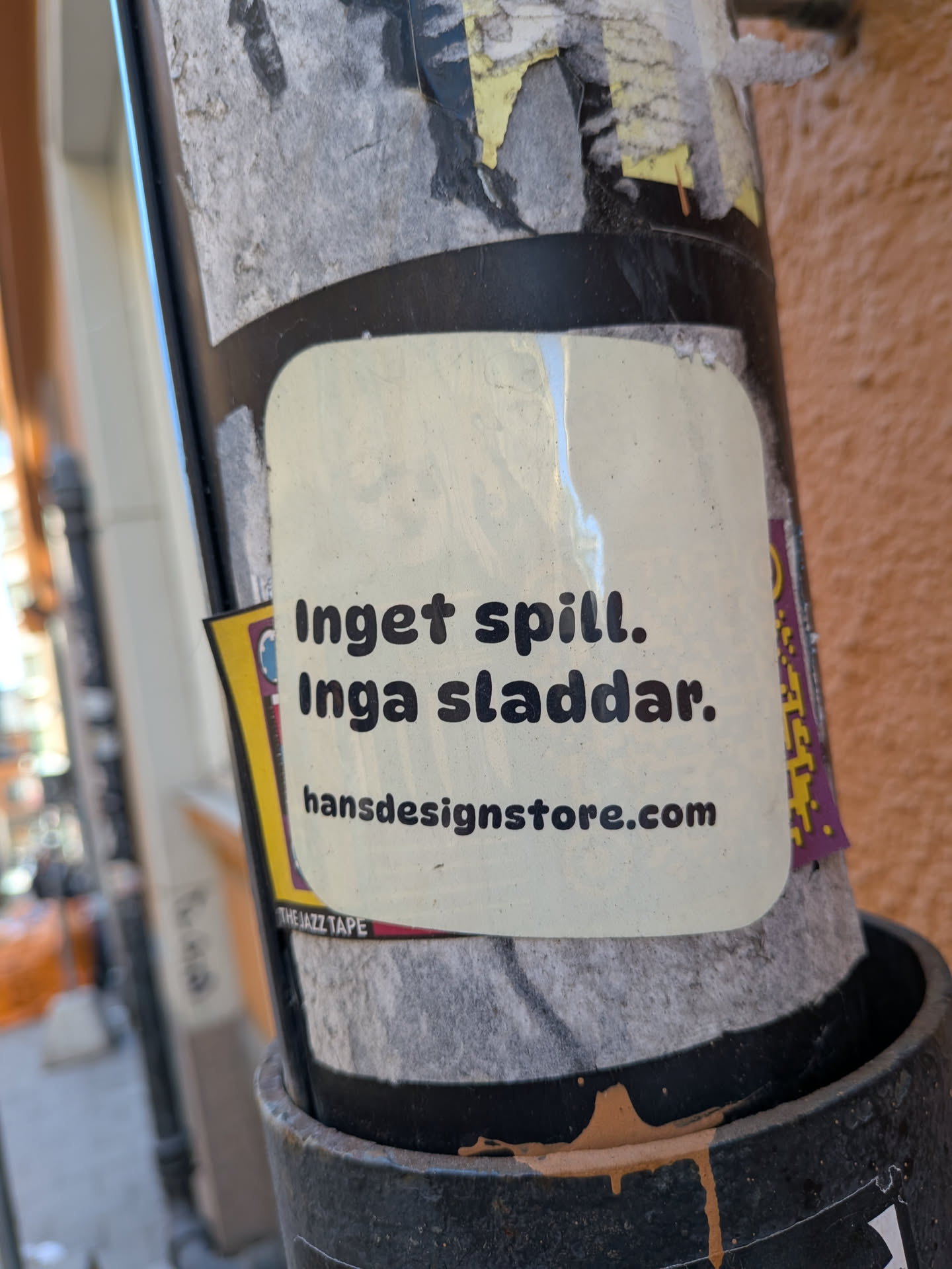 The sticker is rectangular with rounded corners, featuring an off-white or cream background. It displays black, sans-serif text in two lines at the top: 'Inget spill.' and 'Inga sladdar.', followed by a website address 'hansdesignstore.com' in a slightly smaller font at the bottom. The sticker appears slightly worn and dirty, consistent with outdoor placement, and is affixed to a dark pole, partially overlapping other older, torn stickers.
