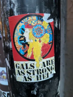 Street sticker The sticker features a circular illustration of a woman's profile with long, flowing blonde hair. She is adorned with an elaborate, colorful headdress featuring geometric patterns and beads in blue, yellow, and red. Her profile is serene, with eyes closed and red lips. The background within the circle is a vibrant mix of blue, orange, and black abstract shapes, possibly representing rays or energy. Below the circular artwork, the text 'GALS ARE STRONG AS HELL' is displayed in bold, black, sans-serif lettering against a white background with a subtle yellow floral pattern. The sticker has a slightly worn appearance with some peeling at the edges, suggesting it has been applied to a surface.