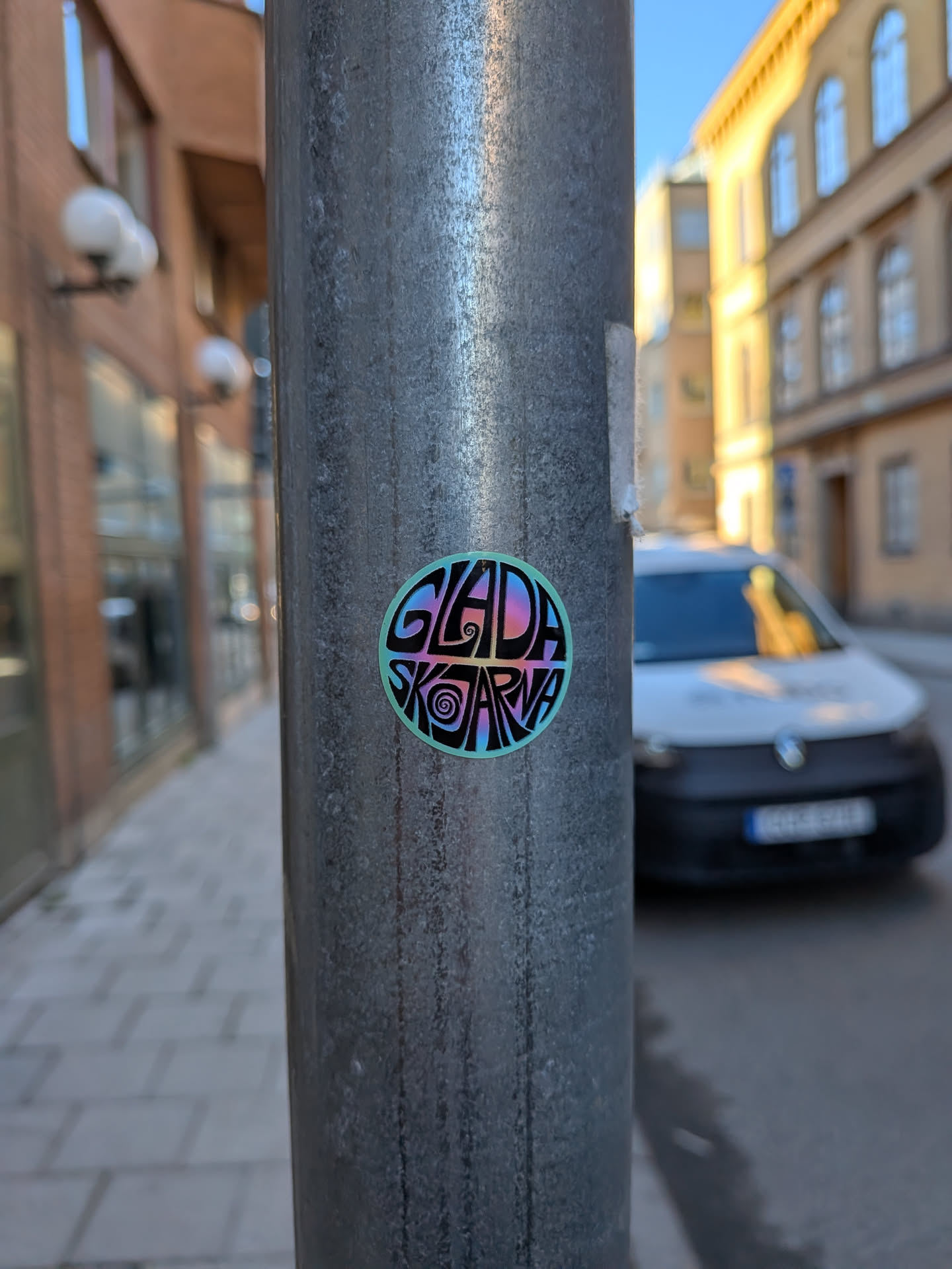 A circular sticker featuring the text 'GLADA SKOJARNA' in a highly stylized, swirly, and somewhat psychedelic font. The letters exhibit an iridescent or holographic effect, displaying a gradient of colors including pink, purple, blue, and green against a solid black background. A thin, light blue-green border encircles the design. The overall appearance is vibrant and playful, resembling a piece of urban street art.
