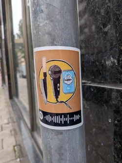 Street sticker Rectangular sticker on a metal pole. Features a yellow circle containing a kitchen knife, a microphone, and a blue 'Tango' can. A black bar at the bottom displays a Spotify code.