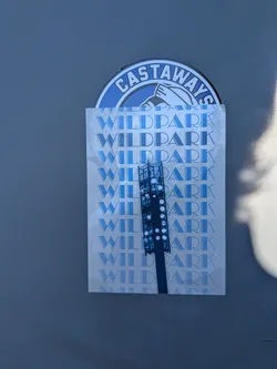Street sticker The sticker is dominated by a repeating pattern of the word 'WILD PARK' in a stylized, art deco-esque blue font against a lighter blue background. Overlaid on this pattern, and positioned centrally, is a dark blue silhouette of a tall communication tower or light pole, detailed with circular lights or emitters. Partially visible at the top is a circular sticker with the word 'CASTAWAYS' in a blue and white emblem, which appears to be a layer underneath or behind the main rectangular sticker.