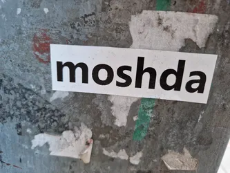 A simple white sticker with the word 'moshda' printed in bold, black, uppercase letters.  It's affixed to a weathered, grey metal surface. 