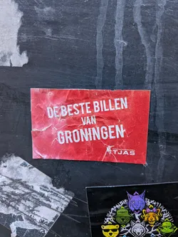 A rectangular red sticker featuring prominent white sans-serif text. The main phrase, "DE BESTE BILLEN VAN GRONINGEN" (The Best Butts of Groningen), is displayed in large, bold capital letters. Below this, in smaller white text, is "Groninger Studenten Schaatsvereniging TJAS" (Groningen Student Skating Association TJAS), accompanied by a small white graphic of what appears to be a stylized figure in motion, possibly skating. The sticker shows significant wear, with numerous wrinkles, creases, and minor tears, particularly along the edges and across its surface, indicating it has been exposed to elements or handled roughly. The red background is faded and discolored in places due to wear.