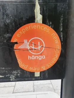 Street sticker An orange round sticker with the word "hänga" in the center and the phrase "New experiences, new moments & new people" written around it. The sticker is slightly damaged and torn.