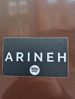 A rectangular black sticker with the name ARINEH written in white capital letters and the Spotify logo below it. The sticker is on a brown surface.