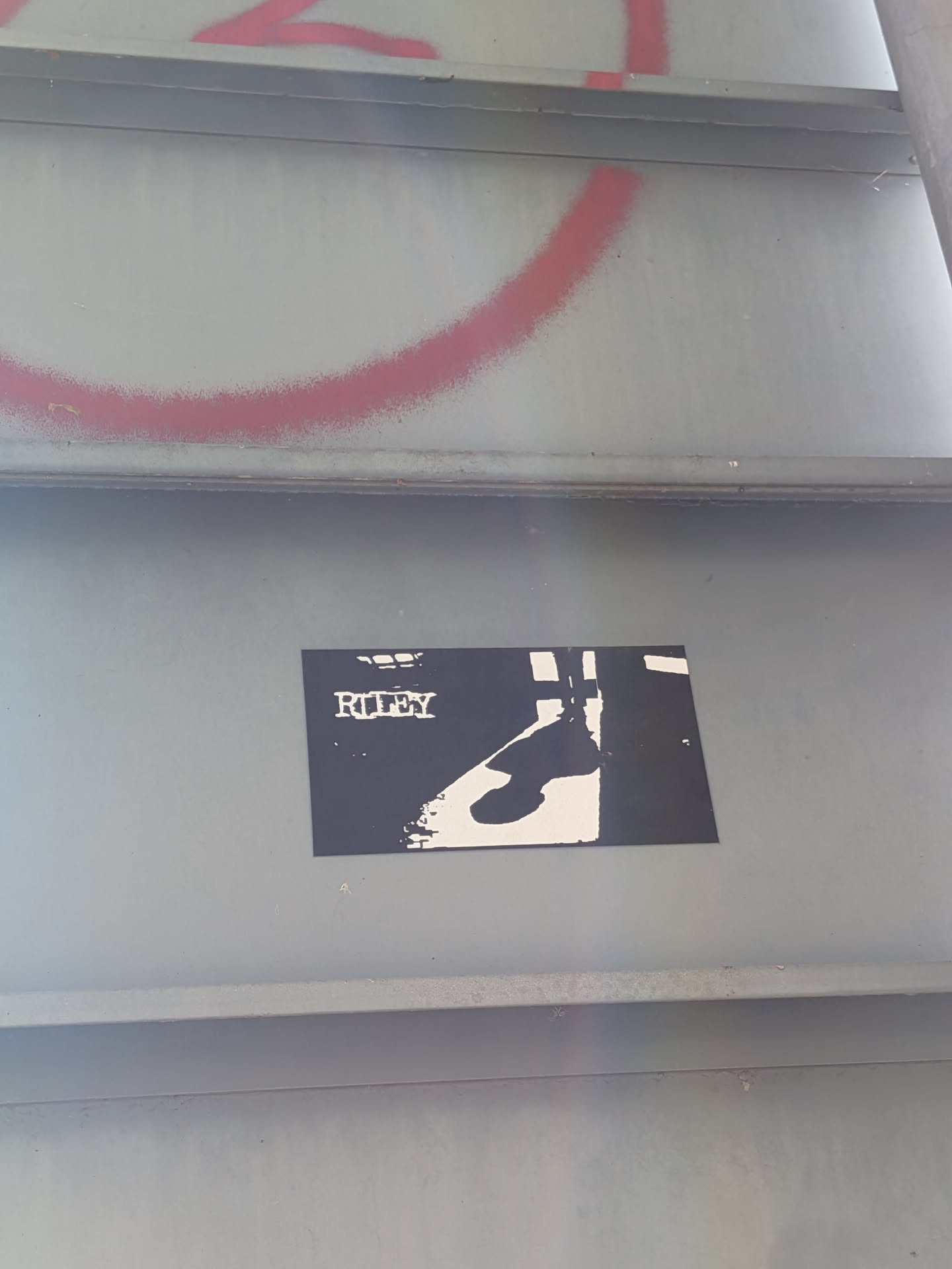 The sticker is rectangular and predominantly black with white elements. At the top, the word 'RILEY' is printed in a distressed, stencil-like white font. Below the text, a silhouette of a person's lower legs and feet is depicted in white against the black background. The silhouette appears to be walking or standing, with one leg slightly forward. The overall aesthetic is minimalist and graphic.
