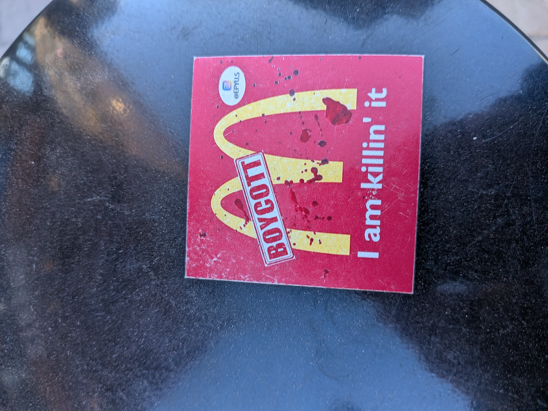 The sticker is rectangular with a vibrant red background. It prominently displays the iconic golden arches (McDonald's 'M' logo) in yellow. Overlaid on the yellow arches and the red background are numerous dark red splatters, resembling blood. A white rectangular banner with a red border is angled diagonally across the 'M', bearing the word "BOYCOTT" in bold red capital letters. To the right of the 'M', in white sans-serif font, is the phrase "I am killin' it". In the top-left corner, there is a small white circular element containing an Instagram logo and the handle "@EPYUS" in black text. The sticker appears slightly worn with some dust and minor scratches.