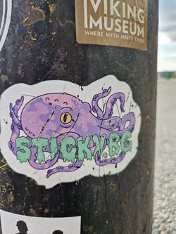 Street sticker A sticker featuring a cartoon octopus in purple and light green colors. The octopus has a friendly expression and its tentacles surround the text "STICKY.BG". The sticker appears to be slightly damaged, showing some tears and scratches.