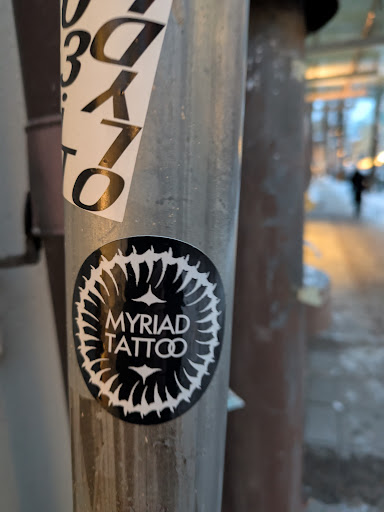 Street sticker Black and white sticker with a circular design and the text 'MYRIAD TATTOO' in the center. The circular design looks like a stylized flower or mandala. The sticker is affixed to a metal pole.