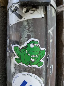 Street sticker The central sticker features a vibrant green cartoon frog, depicted in a sitting position with its front paws slightly raised. The frog has large, expressive white eyes with black pupils looking upwards and to the right, and a wide, friendly smile. Its body is bright green with darker green outlines and a few black spots on its back. Around its neck, the frog wears a white scarf adorned with horizontal black stripes. The entire frog illustration is cut out with a distinct white border, giving it a clean, outlined appearance against the weathered, dark metallic pole it's adhered to.