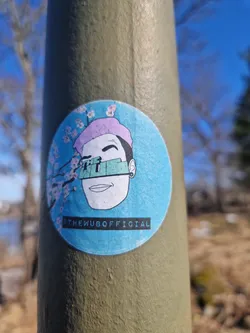 Round sticker featuring a stylized illustration of a person with pink hair, partially obscured by a teal mask or visor.  The background is light blue with pink cherry blossoms. The text '@theWUBofficial' is printed at the bottom.