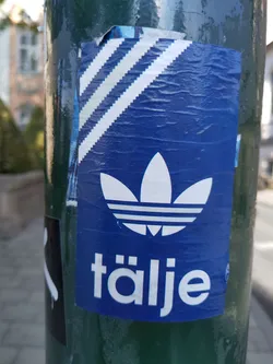Street sticker A blue sticker featuring a stylized Adidas logo and the word "täljje" in white. The sticker is affixed to a green pole.  The sticker appears to be handmade or a fan-made design.