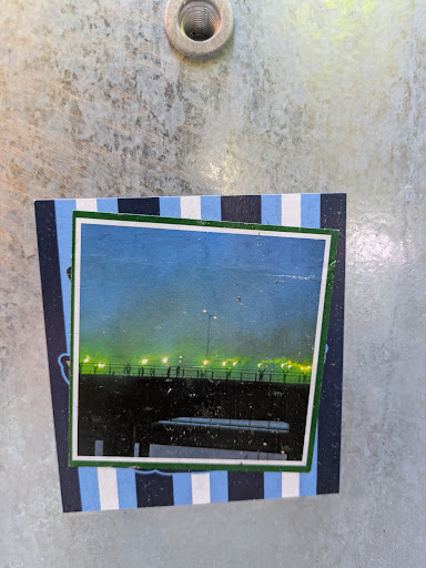 The sticker features a photograph of a bridge at night with lights. The photo is framed in green, and the outer part of the sticker is decorated with blue and white stripes.