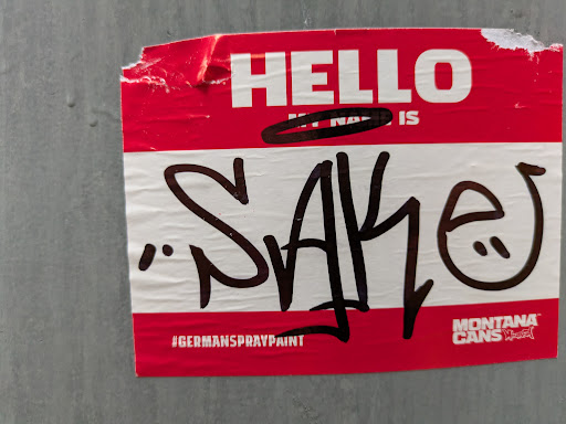 Red and white sticker with graffiti art. The sticker mimics a name tag and includes the text "Hello, my name is" followed by a graffiti tag. At the bottom, it mentions Montana Cans and #germanspraypaint.