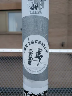 Street sticker The sticker is circular and predominantly black and white. It features a stylized, somewhat abstract depiction of the word 'existence' in a gothic, handwritten font. Below the text, a silhouette of a devilish figure with horns, a tail, and a provocative pose is visible. To the left of the figure is an illustration of what appears to be a microphone or a similar cylindrical object with some detailing. Along the bottom curve of the circle, smaller text reads 'music by even worse people'. The sticker shows signs of wear and tear, with some creasing and discoloration, suggesting it has been applied for some time.