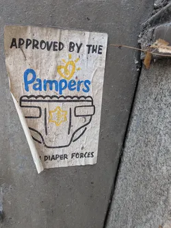 The sticker is a rectangular, off-white piece of paper, heavily soiled and worn, with its bottom left corner peeling away from the dark grey concrete surface. The design features black text at the top reading "APPROVED BY THE". Below this, the word "Pampers" is prominently displayed in a large, stylized blue font, mimicking the brand's logo, complete with a yellow heart-shaped sun icon above it. Beneath the "Pampers" logo is a black outline drawing of a diaper. In the center of the diaper, there is a yellow six-pointed star, which appears to have some faint, unreadable texture or design within it. At the very bottom, partially obscured by dirt and wear, is black text that reads "DIAPER FORCES". The overall appearance is that of a weathered, humorous parody sticker.