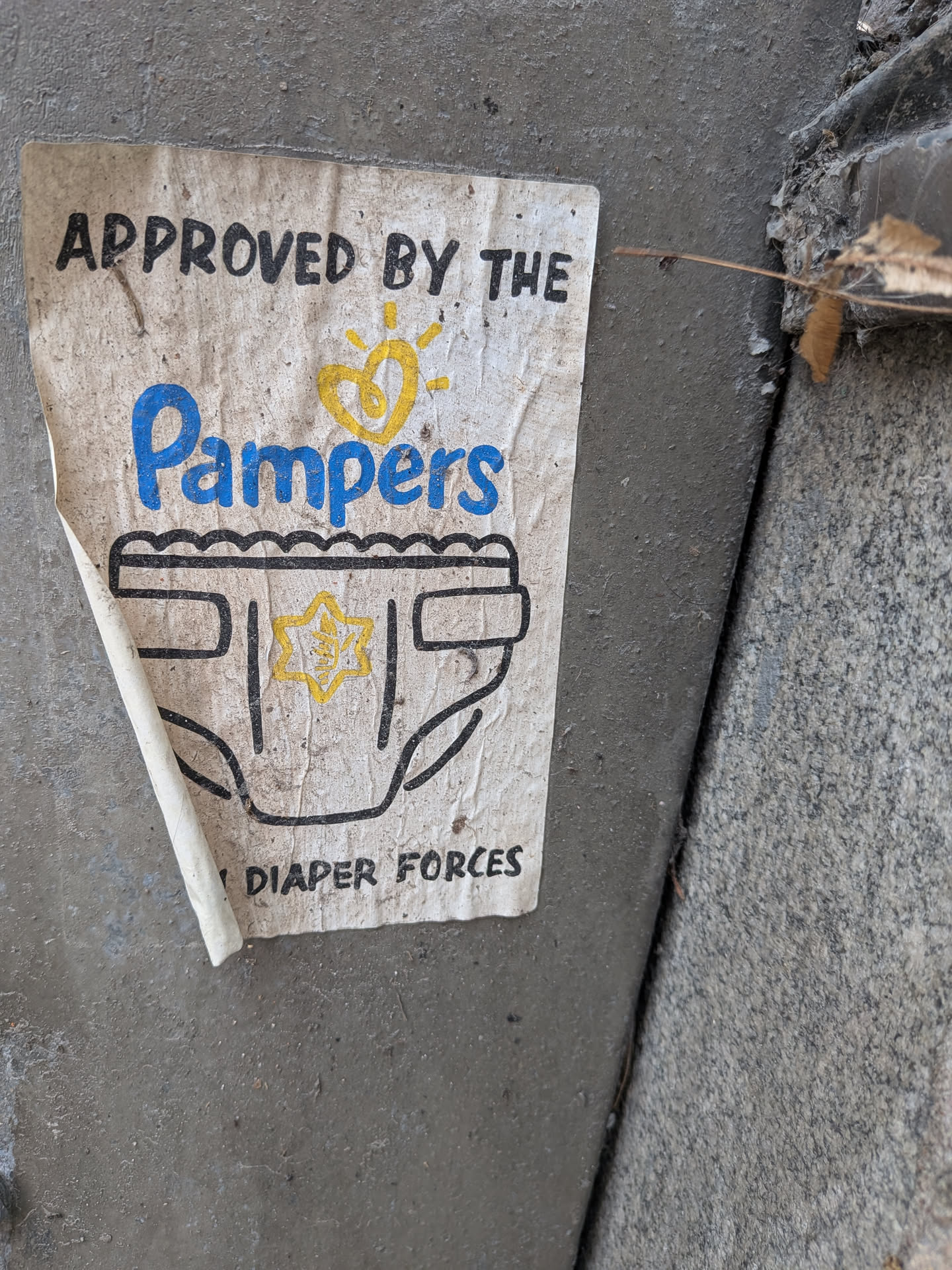 The sticker is a rectangular, off-white piece of paper, heavily soiled and worn, with its bottom left corner peeling away from the dark grey concrete surface. The design features black text at the top reading "APPROVED BY THE". Below this, the word "Pampers" is prominently displayed in a large, stylized blue font, mimicking the brand's logo, complete with a yellow heart-shaped sun icon above it. Beneath the "Pampers" logo is a black outline drawing of a diaper. In the center of the diaper, there is a yellow six-pointed star, which appears to have some faint, unreadable texture or design within it. At the very bottom, partially obscured by dirt and wear, is black text that reads "DIAPER FORCES". The overall appearance is that of a weathered, humorous parody sticker.