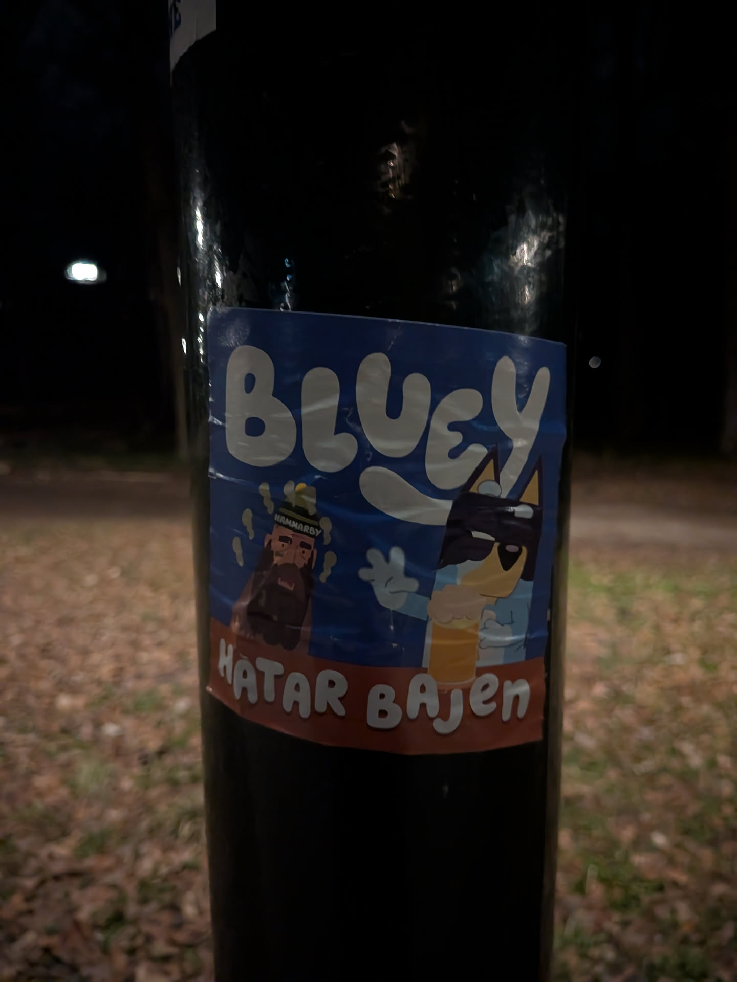 The sticker prominently features the popular blue dog character Bluey, depicted wearing cool black sunglasses and holding a frothy glass of beer. Bluey is shown with a raised hand, as if waving or gesturing. To Bluey's left, a stylized, bearded man with a green and yellow striped beanie, which reads 'HAMMARBY', appears to be sweating or steaming. The upper portion of the sticker has a bright blue background with the word 'BLUEY' in large, bubbly white letters. Below the characters, a red-orange banner spans the width of the sticker, displaying the white text 'HATAR BAJEN'. The sticker has a cartoonish art style and shows visible creases from being applied to the pole.