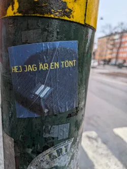 Street sticker A rectangular sticker with a dark blue, textured background, covered in visible water droplets. Bright yellow, uppercase sans-serif text reads 'HEJ JAG ÄR EN TÖNT' across the top. Below the text, a white graphic features two parallel vertical bars enclosed within a diamond shape, outlined in white. The sticker is slightly worn and adheres to a dark green, rough-textured pole.