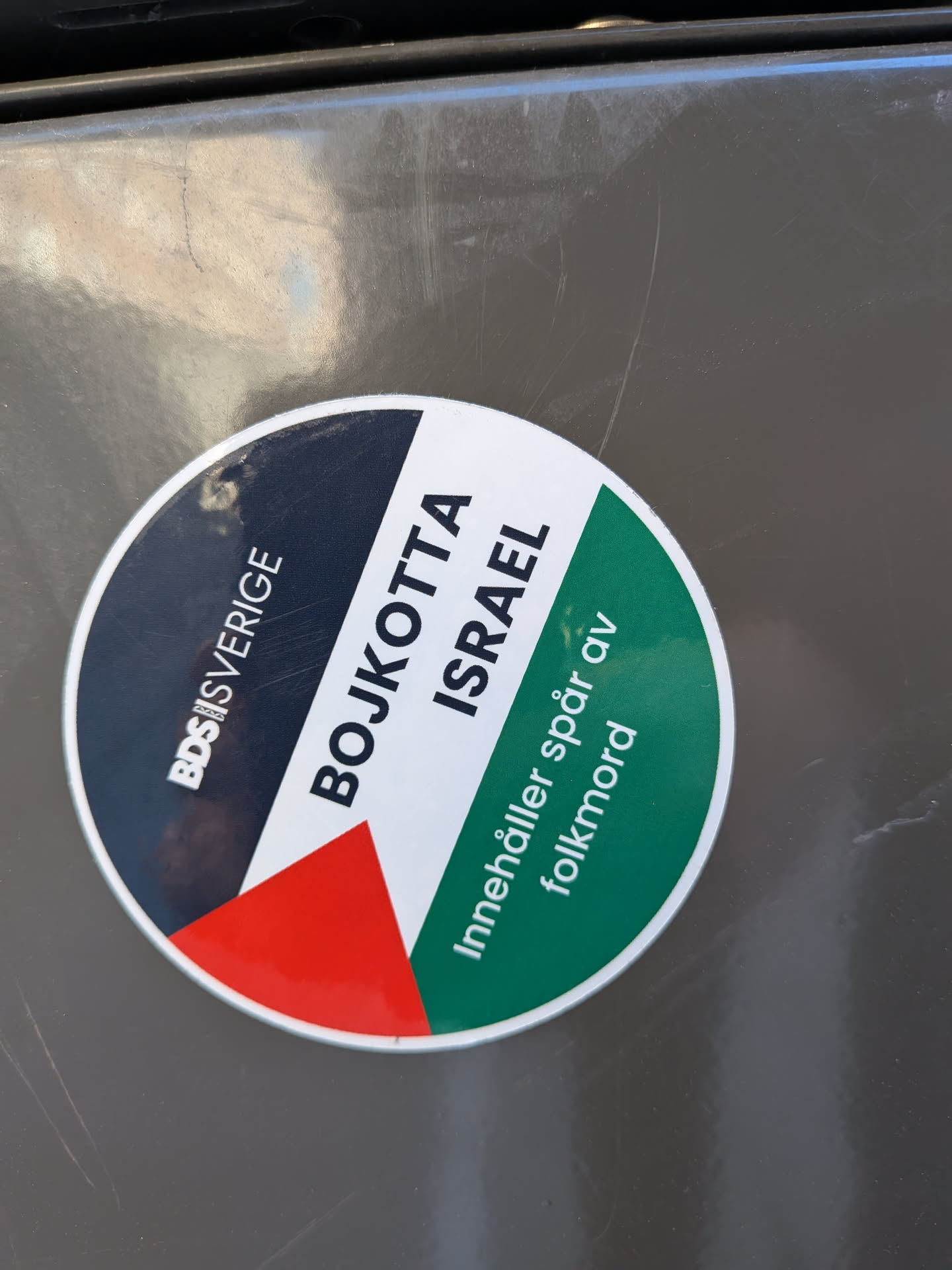 The sticker is circular and divided into sections with different colors and text. The left section is dark blue with white text reading 'BDS|SVERIGE'. Adjacent to this is a segment with the colors of the Palestinian flag: black, white, and red in a triangular shape. The right and top sections are white with large black text that reads 'BOJKOTTA ISRAEL'. The bottom right section is green with white text that reads 'Innehåller spår av folkmord'. The overall design is stark and conveys a strong political message.