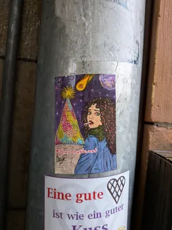 Street sticker The sticker depicts a surreal and cosmic scene. On the right, a woman with voluminous curly brown hair, green eyes, and red lips is shown in profile, looking over her shoulder with a wide-eyed, perhaps startled, expression. She holds a lit cigarette in her mouth and wears a blue jacket over a dark green turtleneck. The background is a deep purple night sky, densely populated with small, sparkling white and pink stars. In the upper left, a bright yellow starburst radiates light. In the upper right, a fiery orange comet with a yellow tail streaks across the sky. Dominating the left side is a large, triangular, pyramid-like structure, intricately patterned with a mosaic of bright colors including yellow, pink, blue, and green, resting on a brown base. The overall style is illustrative, with bold outlines and vibrant colors, giving it a dreamlike or psychedelic quality.
