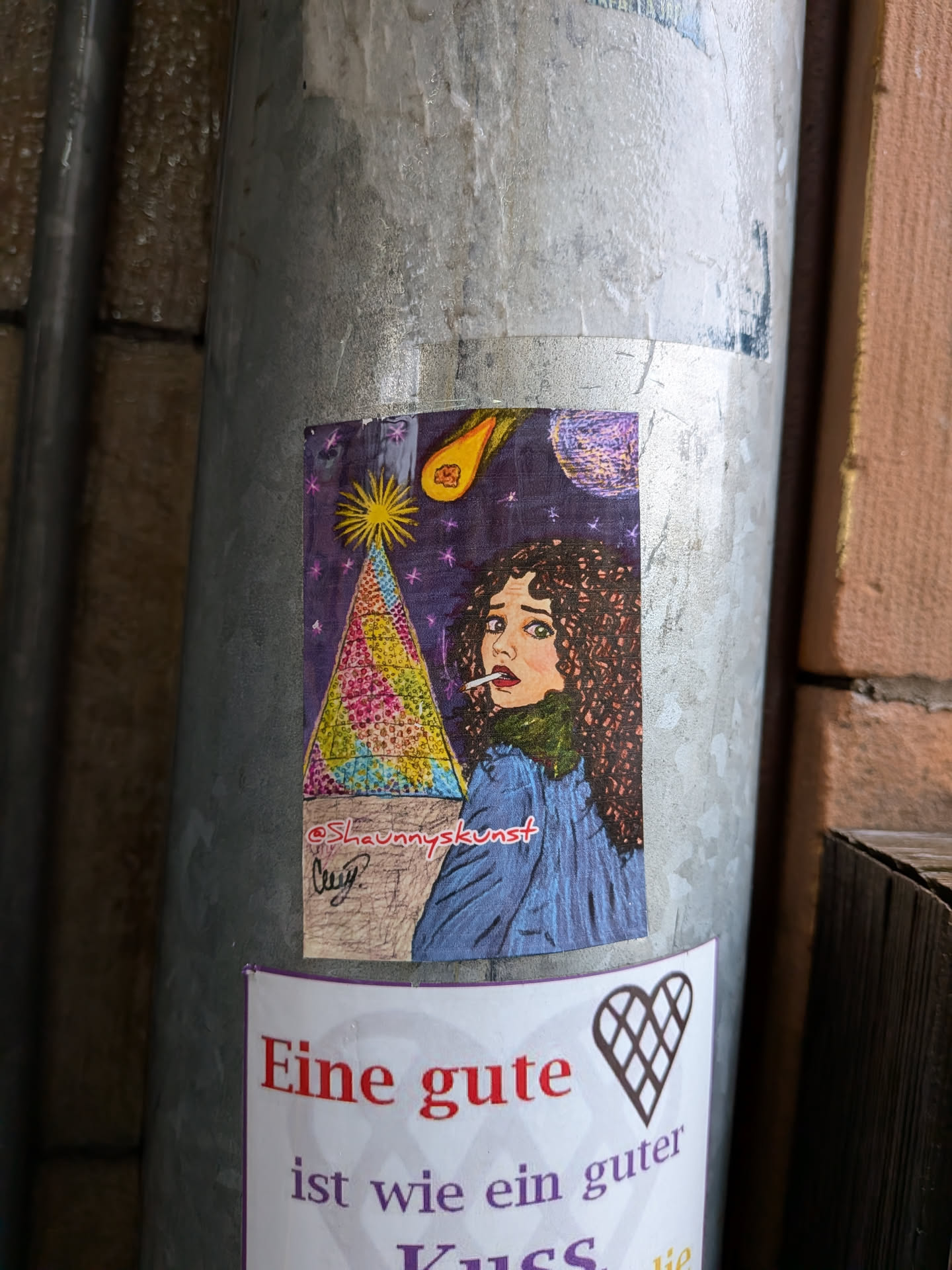 The sticker depicts a surreal and cosmic scene. On the right, a woman with voluminous curly brown hair, green eyes, and red lips is shown in profile, looking over her shoulder with a wide-eyed, perhaps startled, expression. She holds a lit cigarette in her mouth and wears a blue jacket over a dark green turtleneck. The background is a deep purple night sky, densely populated with small, sparkling white and pink stars. In the upper left, a bright yellow starburst radiates light. In the upper right, a fiery orange comet with a yellow tail streaks across the sky. Dominating the left side is a large, triangular, pyramid-like structure, intricately patterned with a mosaic of bright colors including yellow, pink, blue, and green, resting on a brown base. The overall style is illustrative, with bold outlines and vibrant colors, giving it a dreamlike or psychedelic quality.