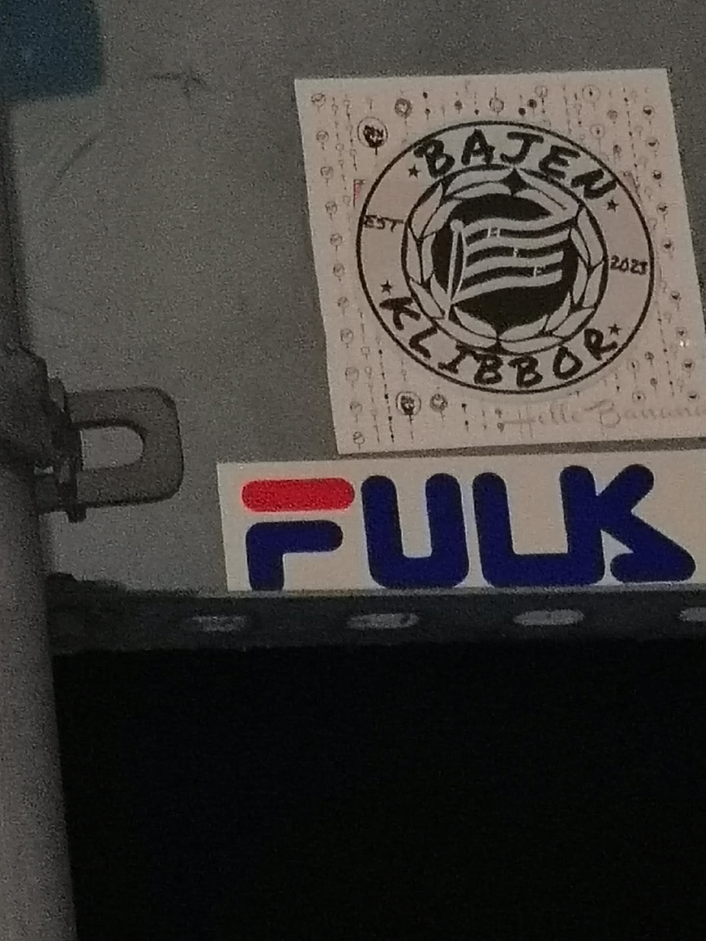 The central sticker is a rectangular white decal featuring a prominent circular emblem in black and white. The emblem depicts a stylized football or globe design with horizontal stripes, possibly representing a flag or a stylized letter. Encircling this design, the text 'BAJEN' is written at the top and 'KLIBBOR' at the bottom, both in a bold, slightly distressed black font. Small text 'EST' is on the left side of the circle and '2023' on the right. The white background of the sticker has a faint, repetitive pattern of small circular or 'Q'-like symbols. Below the main circular design, very faint, handwritten-style text reads 'Helle Banana'.