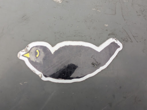 Street sticker A small sticker featuring a simple drawing of a dark-colored bird with a yellow beak. The bird is depicted in a side profile, and its body is slightly curved. The sticker is affixed to a dark grey surface.