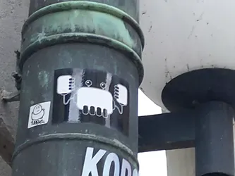 A black and white sticker of a stylized crab with a simple design. The crab is depicted with a rounded body and small, rectangular legs and arms. The sticker has a minimalistic aesthetic and appears to be hand-drawn or digitally created to mimic that style. It's affixed to a dark green metal lamppost.