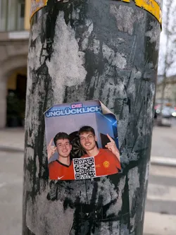 Street sticker The sticker features a photograph of two young men, both smiling and appearing to be friends. They are wearing red shirts with a crest that resembles the Manchester United logo. The man on the left is pointing to his head with his index finger, while the man on the right is also pointing with his index finger. A QR code is prominently placed between them. The background of the photograph appears to be a room with blue walls and some lighting effects. The text 'DIE UNGLÜCKLICH' is visible above the men's heads. The sticker itself is slightly peeling at the top left corner.