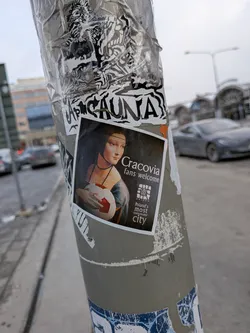 Street sticker The central sticker is a rectangular image featuring a dark-toned, artistic rendition of Leonardo da Vinci's Mona Lisa. She is depicted holding a red and white soccer ball in her arms, with her characteristic enigmatic smile. She wears a dark blue-green dress and a pearl necklace. To the right of the Mona Lisa image, there is white text on a dark background. Below this text, a small, stylized logo for 'Cracovia' is visible, with 'KRAKOW' written underneath it, followed by more text. The entire image is framed by a thin white border.