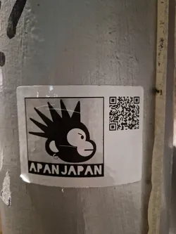 Street sticker A black and white sticker featuring a monkey with spiky hair, a piercing, and an angry expression.  The text 'APANJAPAN' is displayed below the monkey. A QR code is present on the right side of the sticker.