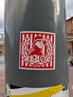 Street sticker The sticker is square with a thin white border. The main background is composed of vertical red and white stripes. Centered on this striped background is a white silhouette of a person, likely a man, wearing a hat, facing slightly to the right. This figure is framed by a white laurel wreath, open at the top. Below the figure, within the wreath, are the stylized numbers "1910" in white.