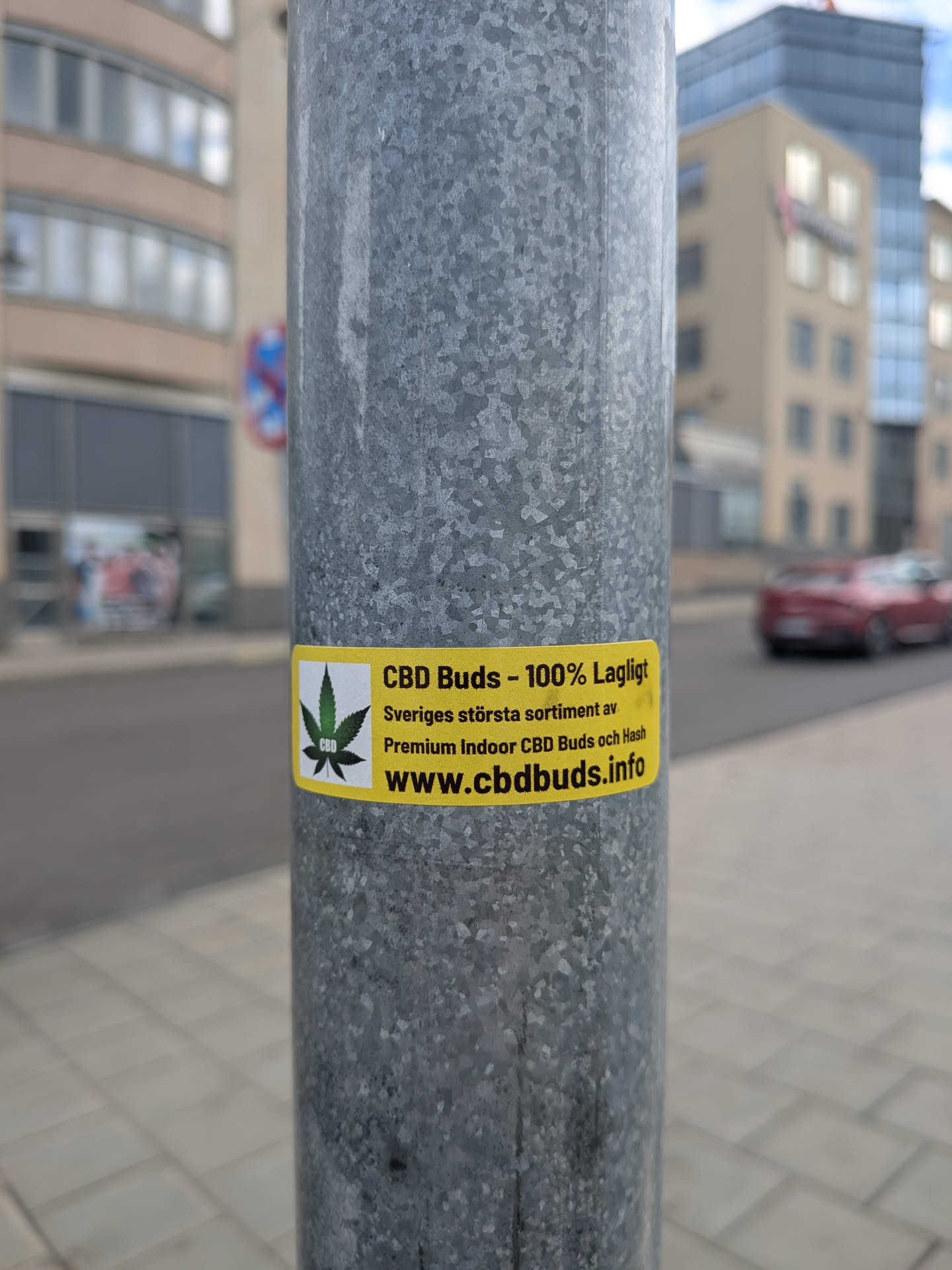 A rectangular yellow sticker affixed to a metal pole. The left side features a green cannabis leaf icon on a white square. The right side contains Swedish text advertising CBD buds and a website URL.