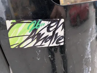 Street sticker The sticker is rectangular with slightly rounded corners, adhered to a dark, worn surface. Its background is primarily white, with a vibrant, light green, vertically scribbled section on the left side, resembling marker or crayon strokes. Overlaid on this background is prominent, stylized text in thick, black brush strokes, which appears to read 'El Andre'. The lettering has an energetic, raw, and graffiti-like quality. In the upper left portion, partially overlapping both the green scribbles and the black text, are the numbers '26"' written in a brighter, slightly darker green marker, also in a casual, handwritten style. The overall aesthetic is a blend of raw street art and custom tagging.