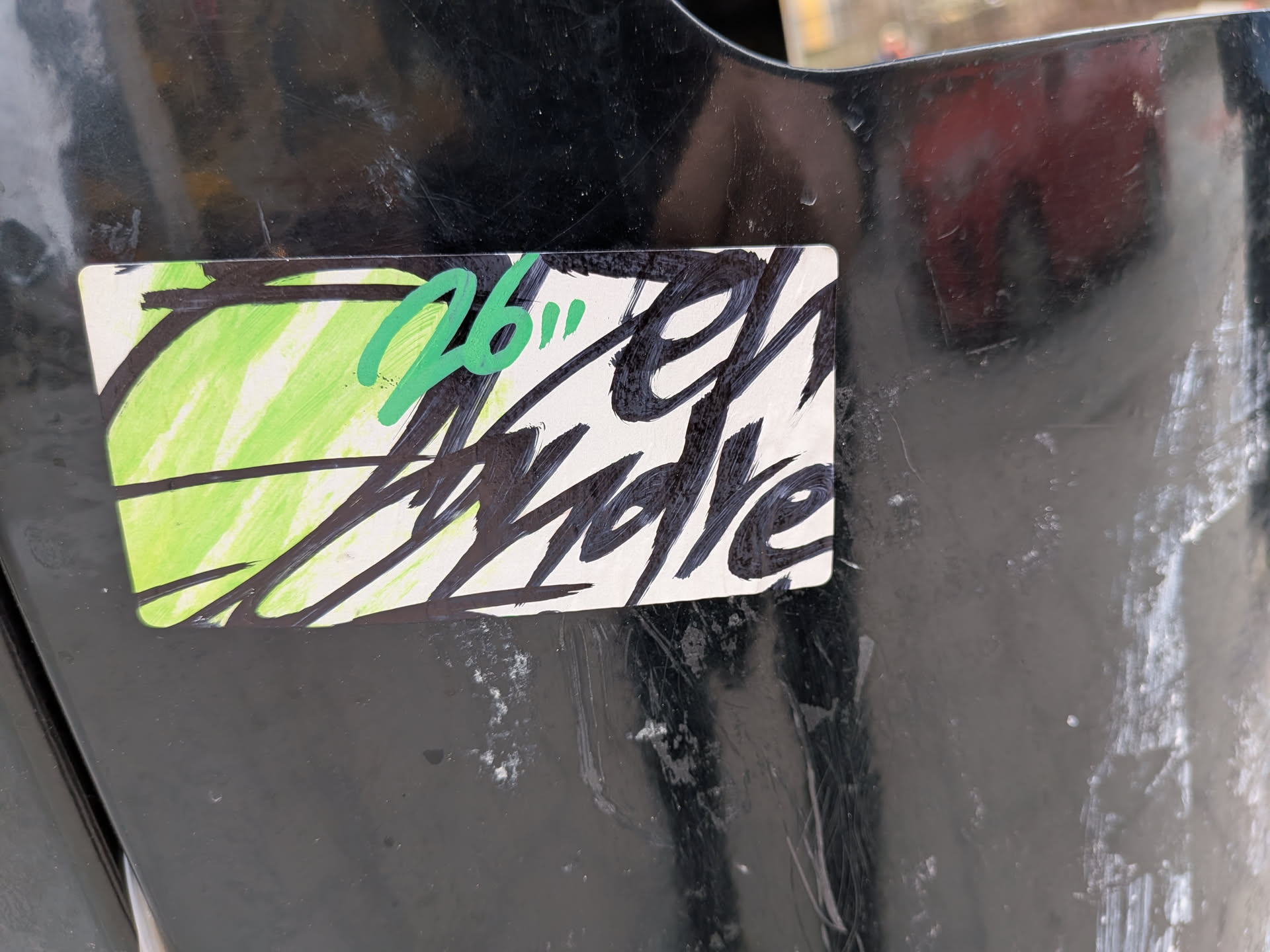 The sticker is rectangular with slightly rounded corners, adhered to a dark, worn surface. Its background is primarily white, with a vibrant, light green, vertically scribbled section on the left side, resembling marker or crayon strokes. Overlaid on this background is prominent, stylized text in thick, black brush strokes, which appears to read 'El Andre'. The lettering has an energetic, raw, and graffiti-like quality. In the upper left portion, partially overlapping both the green scribbles and the black text, are the numbers '26"' written in a brighter, slightly darker green marker, also in a casual, handwritten style. The overall aesthetic is a blend of raw street art and custom tagging.