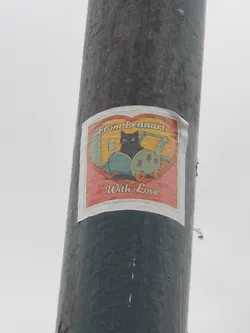 Street sticker The sticker is roughly square with a white border, featuring a large red heart shape as its central frame. Inside the heart, a stylized black cat with bright green eyes sits comfortably in a light blue/teal baby stroller. The stroller has orange wheels and is adorned with small red roses on its side. The background within the heart is a warm yellow, showing a white electrical outlet on the wall behind the cat. Above the cat, within the heart, the text "From Lennart" is written in a playful, slightly retro font. Below the cat, also within the heart, the text "With Love" is displayed in a similar style. The overall aesthetic is cartoonish and evokes a sense of warmth and affection.