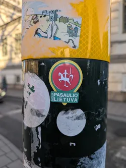 Street sticker A circular sticker featuring the white Vytis emblem – a knight on a rearing white horse, holding a silver sword and shield – set against a vibrant red background. The central red circle is outlined with thin white, green, and yellow borders. Below the emblem, a green banner-shaped element contains the white text 'MISIJA PASAULIO LIETUVA'. The sticker is affixed to a black pole, showing minor signs of wear.
