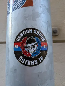 Street sticker A circular sticker with a dark brown background, featuring a central white skull wearing a black fedora with a white band. The skull is framed by two laurel wreaths, one red on the left and one blue on the right. Behind the skull and laurels, there's a horizontal stripe, red on the left and blue on the right. White text arcs along the top edge reading "SEKTION SÖDRA" and along the bottom edge reading "ÖSTERS IF". The numbers "20" and "21" are placed on the red and blue stripes respectively, to the left and right of the laurel wreaths.