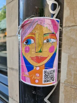 Street sticker A vibrant sticker featuring a stylized female face with bold colors and abstract shapes. The artist's name, Claire View, is subtly printed at the bottom, along with a QR code. The face has pink hair, large colorful eyes and pink circles on the cheeks. 