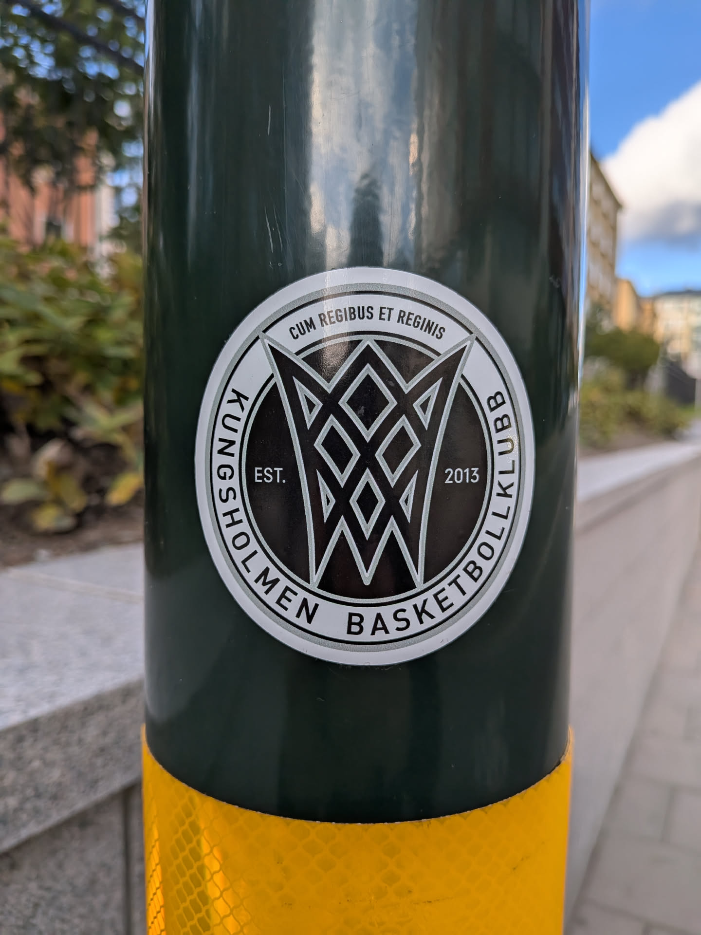 Street sticker The sticker is circular with a white border. The central design features a stylized, abstract representation of a basketball hoop and net, rendered in black and white with sharp geometric lines. Inside the central design, the text 'EST.' is visible on the left, and '2013' is visible on the right. The outer ring of the sticker contains text in black capital letters on a white background, with a thin black outline. The text reads 'KUNGSHOLMEN BASKETBOLLKLUBB' along the bottom arc and 'CUM REGIBUS ET REGINIS' along the top arc.