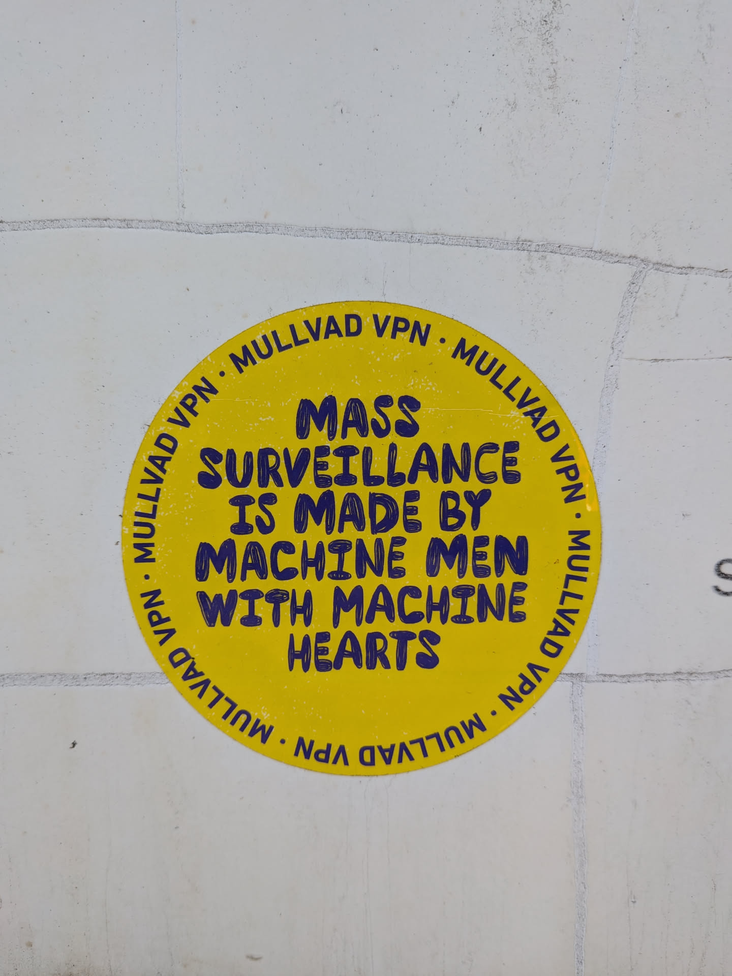A circular yellow sticker with a textured, slightly worn surface. The text is printed in a dark blue, slightly irregular, blocky font. The outer edge of the sticker features the text 'MULLVAD VPN' repeated multiple times, separated by dots. The inner text reads 'MASS SURVEILLANCE IS MADE BY MACHINE MEN WITH MACHINE HEARTS'.