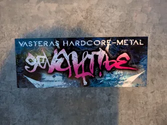 Street sticker The sticker features a vibrant, stylized graphic against a dark, textured background with streaks of blue and white. Prominently displayed is a band logo in a graffiti-like font, with gradient colors transitioning from white to pink and bright magenta. The text 'VASTERAS HARDCORE-METAL' is printed in white, sans-serif, stencil-like font across the top of the sticker. The overall aesthetic suggests a connection to the music scene, likely hardcore or metal.