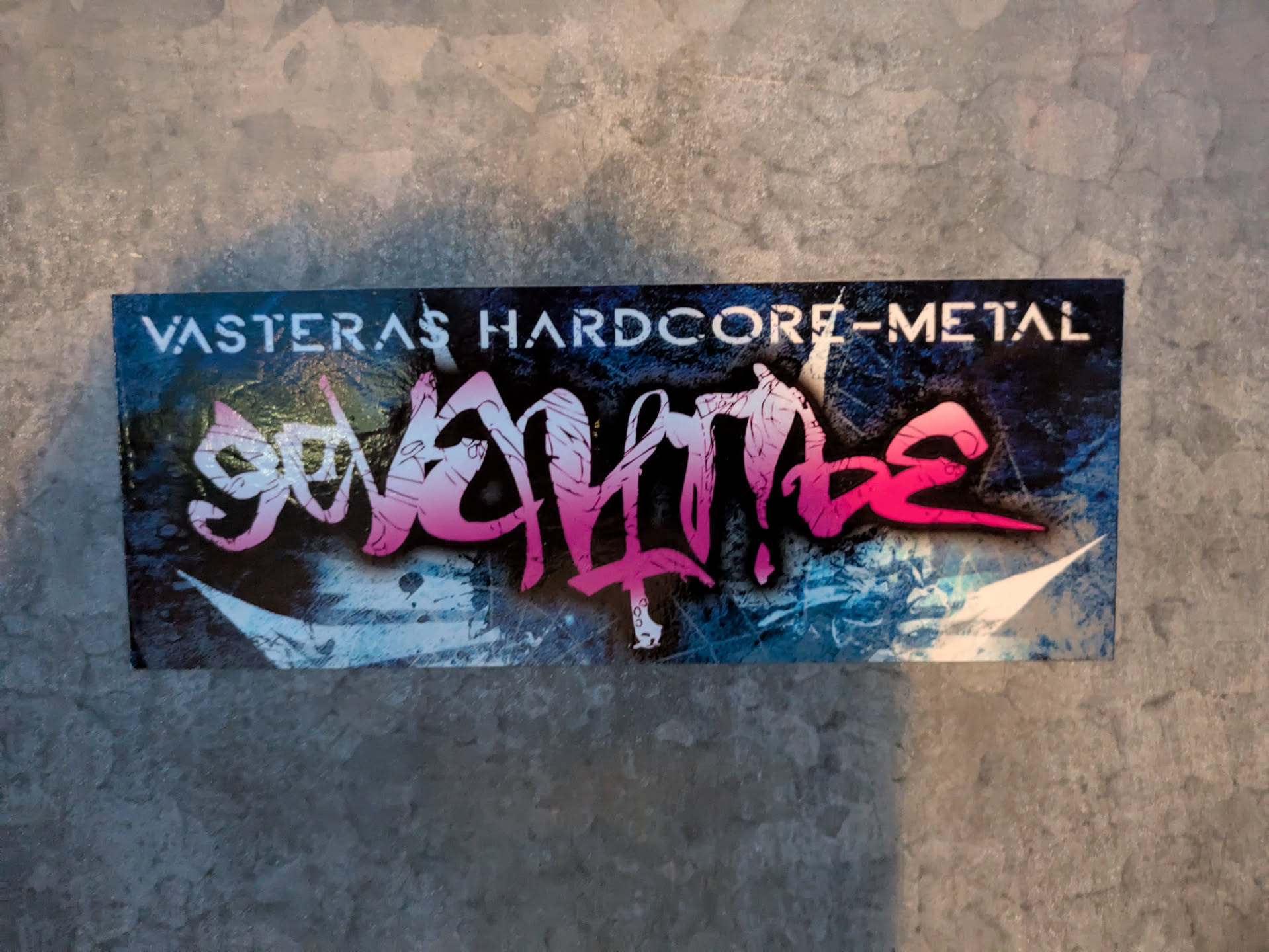 A rectangular sticker featuring a dark, distressed background with splatters of blue and white, resembling an abstract, icy texture or aged concrete. Dominating the center is the band name 'Senntia' rendered in a bold, stylized, almost jagged graffiti-like font, transitioning in color from vibrant pink on the right to a lighter magenta and white on the left. Above this, in a clear, white stencil-like font, are the words 'VASTERAS HARDCORE-METAL'. The overall aesthetic is gritty and energetic, fitting for the music genre.
