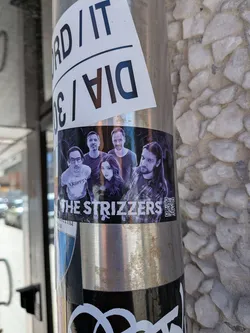 Street sticker A horizontally oriented rectangular sticker adhered to a metallic pole, showcasing a group portrait of five individuals, presumably the band members of 'THE STRIZZERS'. The overall aesthetic is dark and moody, with the figures set against a black background with subtle purple and blue smoky effects. From left to right, the members include a man with glasses wearing a white 'TRIPPIN'' t-shirt, two men with dark t-shirts, a woman with long dark hair in the center, and a man with long hair and a beard wearing a plaid shirt on the right. Below the portrait, the band's name 'THE STRIZZERS' is prominently displayed in a distressed white font. A black and white QR code is located to the right of the band's name.