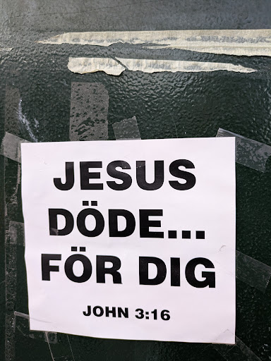 A sticker, likely religious in nature, is affixed to a green surface with tape. The sticker reads "JESUS DÖDE... FÖR DIG JOHN 3:16", which translates to "Jesus Died For You John 3:16". The text is printed in a bold sans-serif font. The sticker itself is white.