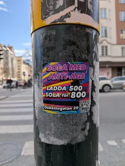The sticker is rectangular with slightly rounded corners, featuring a highly vibrant and abstract background composed of horizontal wavy stripes in shades of pink, purple, yellow, and blue, creating a dynamic, almost retro-futuristic aesthetic. Bold, white text with a purple outline reads "SOLA MED ANTI-AGE" at the top. Below this, in a similar style, it states "LADDA 500 SOLA för 800", indicating a promotional offer. At the bottom, smaller white text with a blue outline specifies the address "Döbelnsgatan 39". A black and white QR code is prominently displayed in the bottom right corner. The sticker is affixed to a dark, textured pole, with some older, torn sticker remnants visible around it.
