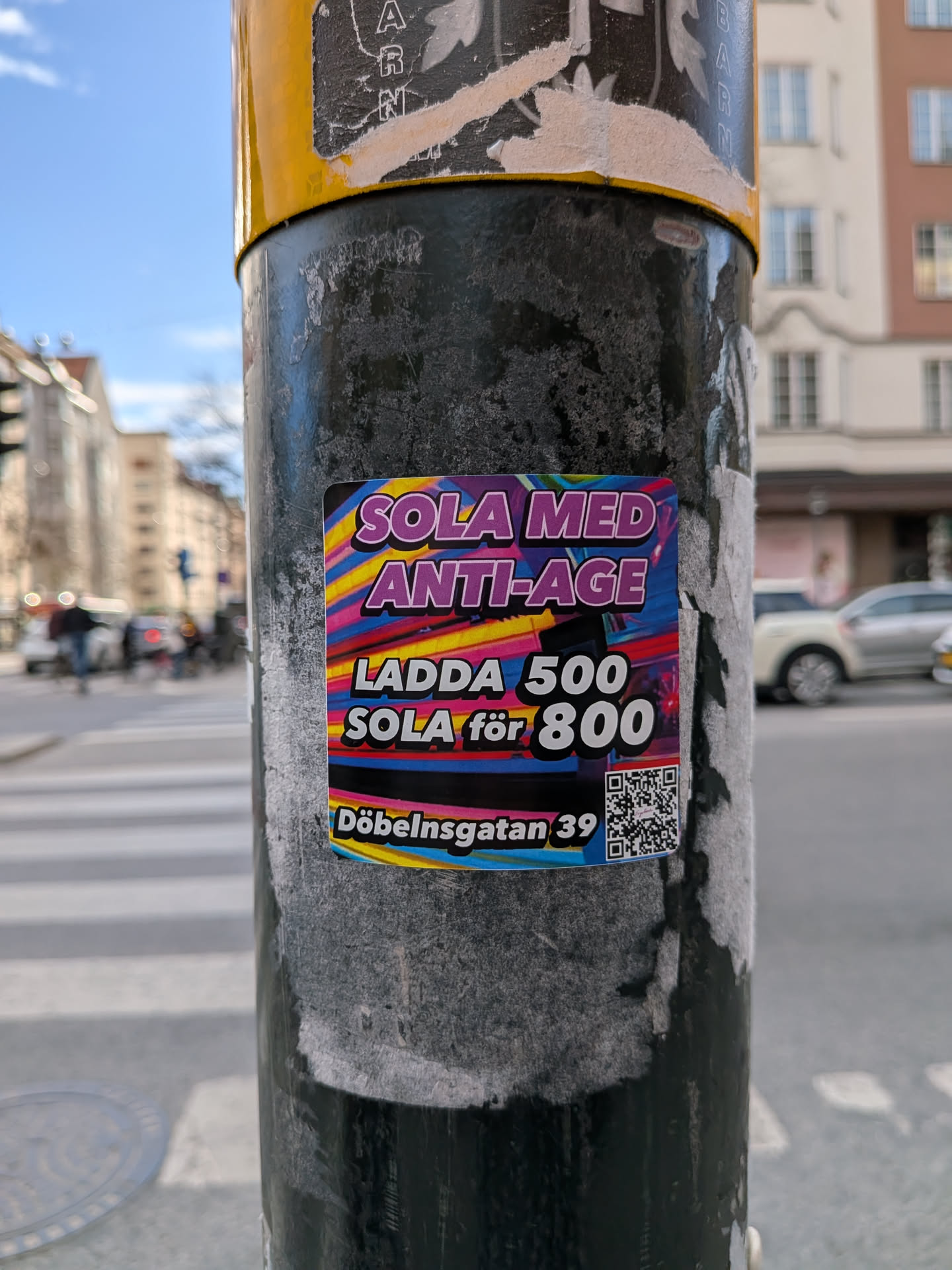 The sticker is rectangular with slightly rounded corners, featuring a highly vibrant and abstract background composed of horizontal wavy stripes in shades of pink, purple, yellow, and blue, creating a dynamic, almost retro-futuristic aesthetic. Bold, white text with a purple outline reads "SOLA MED ANTI-AGE" at the top. Below this, in a similar style, it states "LADDA 500 SOLA för 800", indicating a promotional offer. At the bottom, smaller white text with a blue outline specifies the address "Döbelnsgatan 39". A black and white QR code is prominently displayed in the bottom right corner. The sticker is affixed to a dark, textured pole, with some older, torn sticker remnants visible around it.