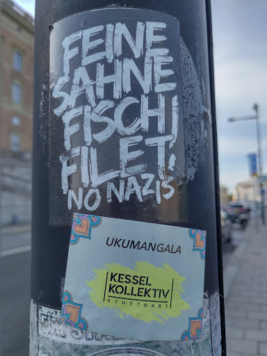 Street sticker Two stickers are attached to a pole. The top sticker features the text "Feine Sahne Fischfilet, No Nazis" written in a stylized graffiti font. The bottom sticker is smaller and features the text "UKUMANGALA Kessel Kollektiv Stuttgart". The design of the second sticker includes decorative corner elements.